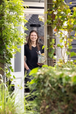 Yara poseert in een deuropening, tussen het groen. Yara is een jonge vrouw met bruin haar, ze draagt een zwart t-shirt. Ze lacht breed. In haar hand houdt ze een pot met schildersspullen.