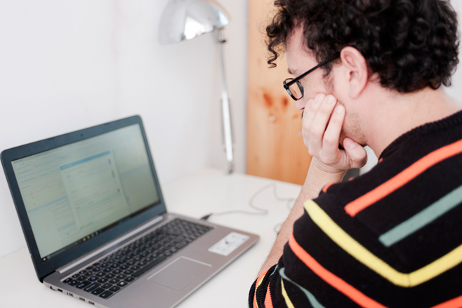 Een laptop staat op een bureau. Erachter zit een man in een gestreepte trui. Hij heeft krullend haar, draagt een bril. Zijn hoofd steunt op zijn hand.