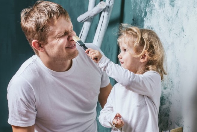 Vader zit met dochter bij een ladder. Dochter strijkt met kwast over zijn neus.