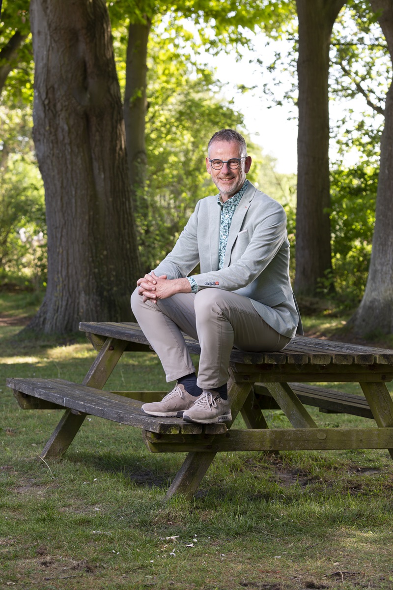 Ansfried Snijders poseert op een picknicktafel. Hij draagt een grijs pak met een blauw overhemd. Hij heeft kort grijs haar, een bril en lacht.
