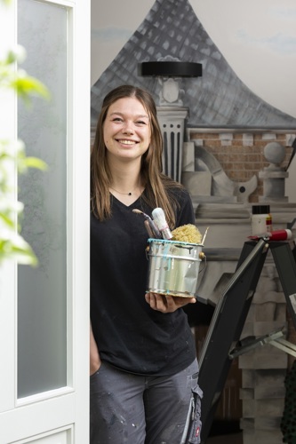 Yara poseert in de deuropening van een B&B. Ze heeft lang bruin haar, lacht haar tanden bloot en draagt een zwart t-shirt. In haar hand houdt ze een pot met kwasten.