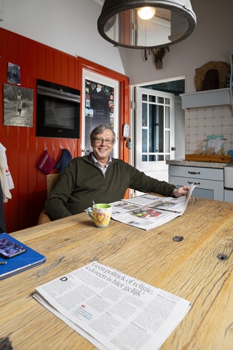 Frans Boetzkes zit aan de keukentafel. Op de keukentafel ligt een krant open. Hij lacht.