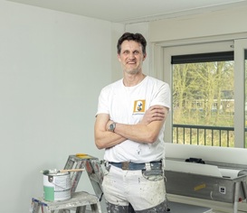 Jan van Loosen poseert voor een raam. Hij heeft zijn armen over elkaar, witte kleding aan en lacht.